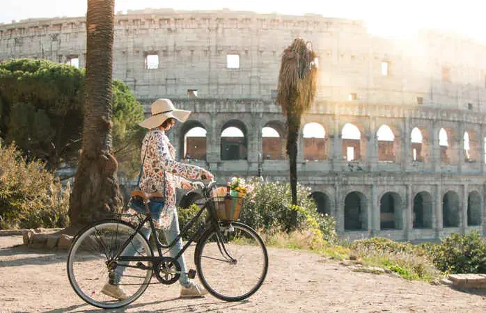 Beautiful young woman in colorful fashion dress walking alone on hill with her bike in front of colosseum in Rome at sunset with trees. Attractive tourist girl with straw hat. Lens Flare.